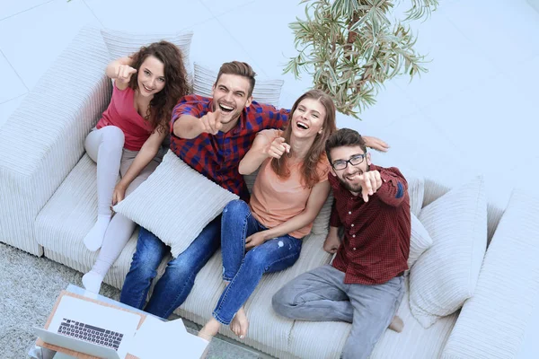 Ομάδα γέλιο φίλοι κάθονται στον καναπέ και προβολή δείκτη στην κάμερα. — Φωτογραφία Αρχείου