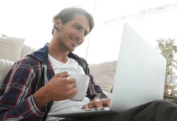 Jovem com laptop segurando uma xícara sentada no chão perto do sofá — Fotografia de Stock