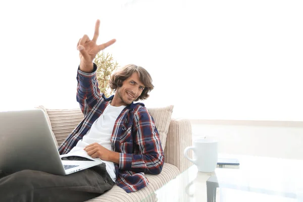 Chico feliz con el ordenador portátil sentado en el sofá y mostrando su mano un gesto ganador —  Fotos de Stock