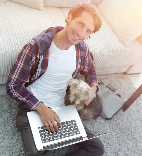 Stilig kille med laptop kramar hans hund och sitter nära soffan — Stockfoto