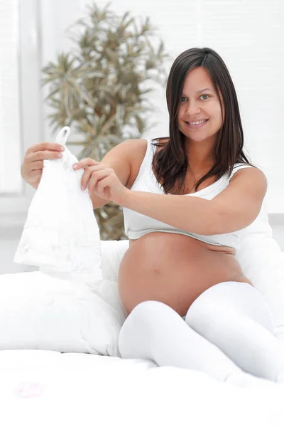Mulher grávida considerando camisa das crianças, sentado na cama . — Fotografia de Stock