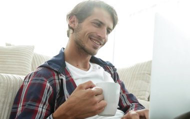 genç adam kanepede yakın katta oturan bir fincan tutan dizüstü bilgisayar ile