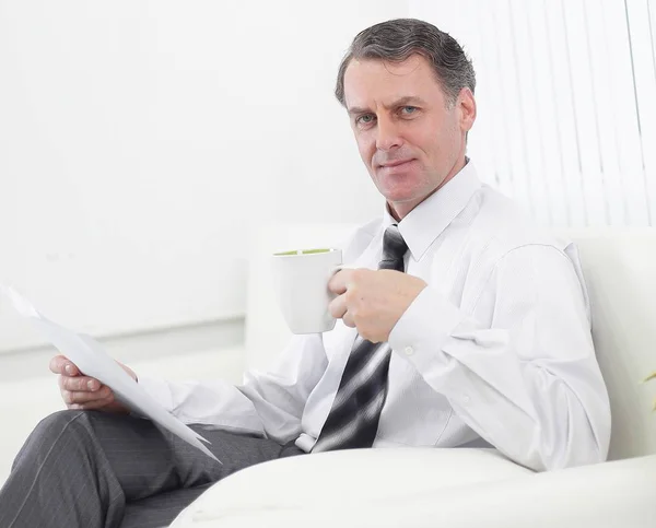 Confident businessman drinking tea and reading a document,sitting in a chair in the hotel room — Stock Photo, Image