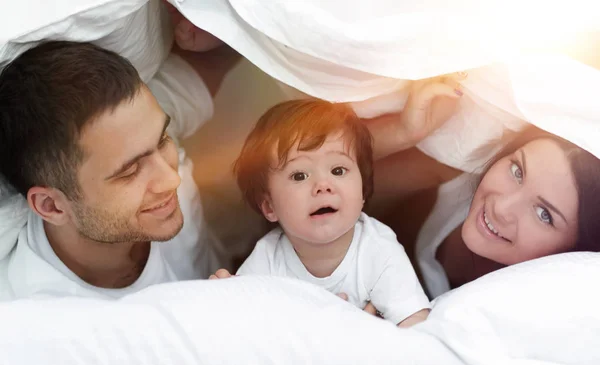 Familia feliz posando bajo un edredón mientras mira a la cámara —  Fotos de Stock