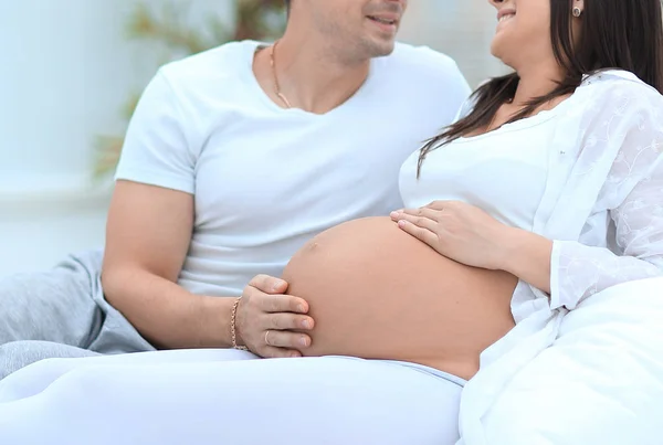 Mulher grávida feliz e parceiro sentado na cama com as mãos tocando barriga — Fotografia de Stock