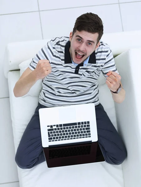 Vue du haut. jubilant jeune homme avec ordinateur portable assis sur le canapé — Photo