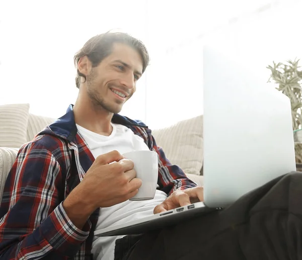 Jovem com laptop segurando uma xícara sentada no chão perto do sofá — Fotografia de Stock