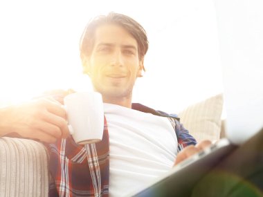 genç adam kanepede yakın katta oturan bir fincan tutan dizüstü bilgisayar ile