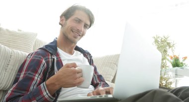 genç adam kanepede yakın katta oturan bir fincan tutan dizüstü bilgisayar ile