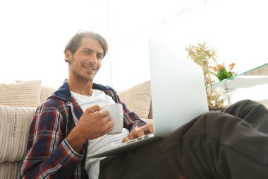 genç adam kanepede yakın katta oturan bir fincan tutan dizüstü bilgisayar ile