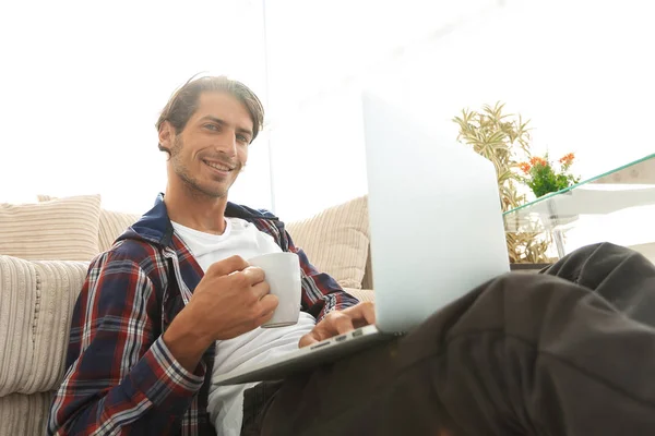 Jovem com laptop segurando uma xícara sentada no chão perto do sofá — Fotografia de Stock