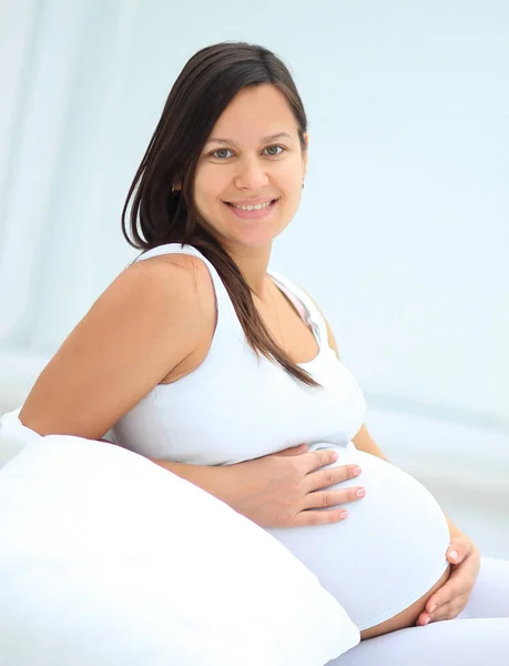 Beautiful pregnant woman sitting at sofa and keeping hand on belly. — Stock Photo, Image