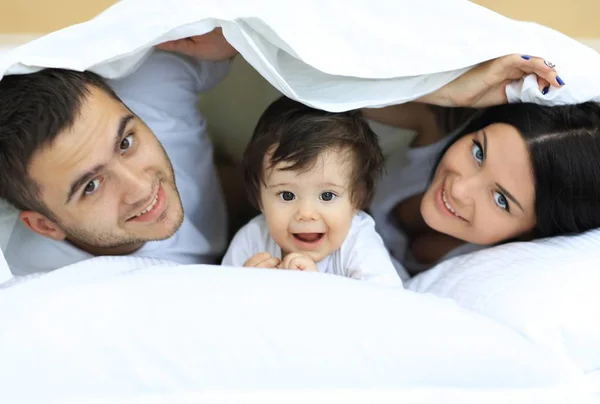 Familia feliz posando bajo un edredón mientras mira a la cámara —  Fotos de Stock
