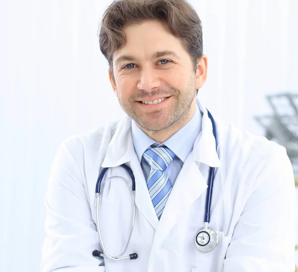 Guapo joven médico en bata blanca está mirando a la cámara y sonriendo mientras está de pie en la oficina — Foto de Stock