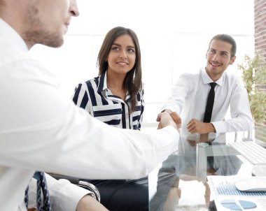 Erkekler masada otururken el sıkışıyor ve gülümsüyor.