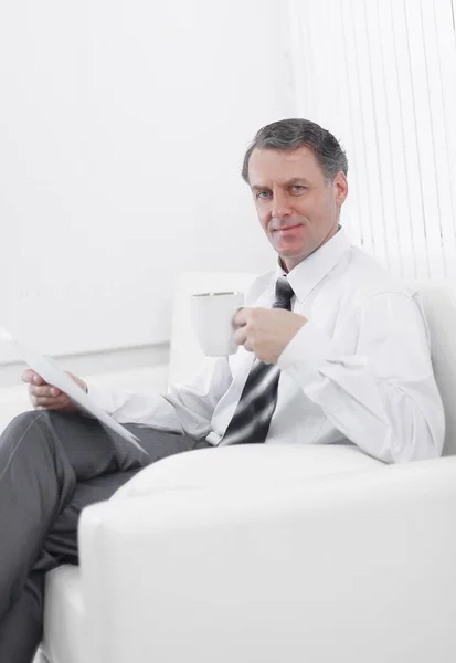 Hombre de negocios confiado bebiendo té y leyendo un documento, sentado en una silla en la habitación del hotel — Foto de Stock