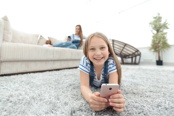 Nettes kleines Mädchen mit Smartphone auf dem Teppich im Wohnzimmer liegend — Stockfoto