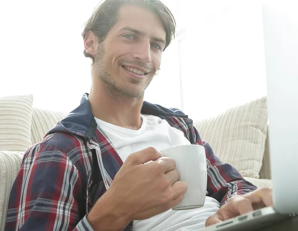 Молодий чоловік з ноутбуком тримає чашку, що сидить на підлозі біля дивана — стокове фото