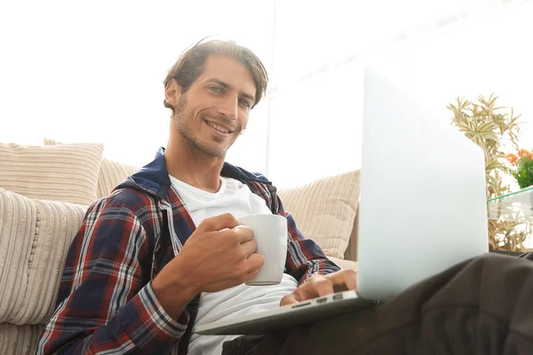 Jovem com laptop segurando uma xícara sentada no chão perto do sofá — Fotografia de Stock