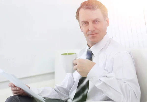 Hombre de negocios confiado bebiendo té y leyendo un documento, sentado en una silla en la habitación del hotel —  Fotos de Stock