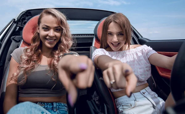 Duas jovens mulheres felizes sentadas em um carro e apontando para você . — Fotografia de Stock