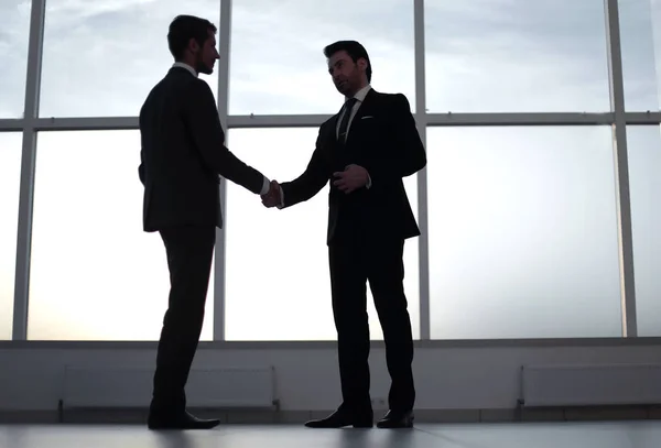 Businessmen shake hands, standing near a large office window — Stock Photo, Image