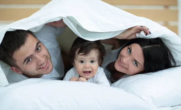 Familia feliz posando bajo un edredón mientras mira a la cámara —  Fotos de Stock