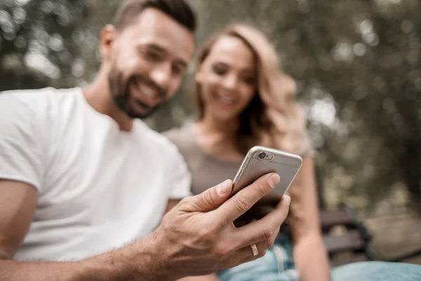 Jovem casal assiste a um vídeo em um smartphone sentado em um parque b — Fotografia de Stock