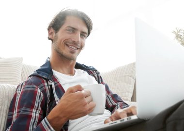 genç adam kanepede yakın katta oturan bir fincan tutan dizüstü bilgisayar ile