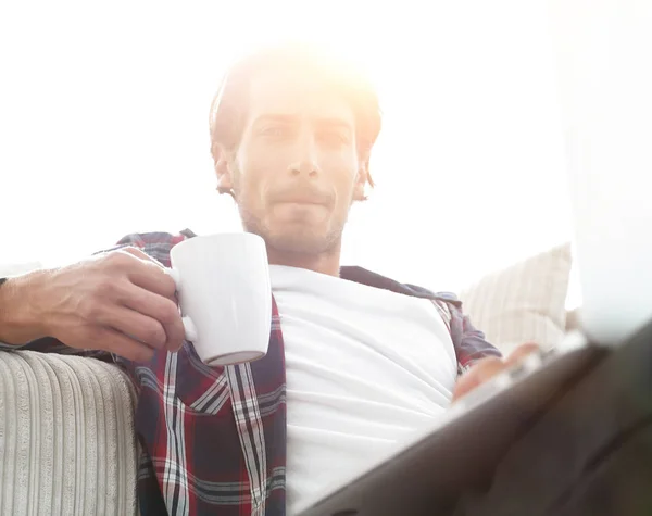 Joven con portátil sosteniendo una taza sentada en el suelo cerca del sofá — Foto de Stock