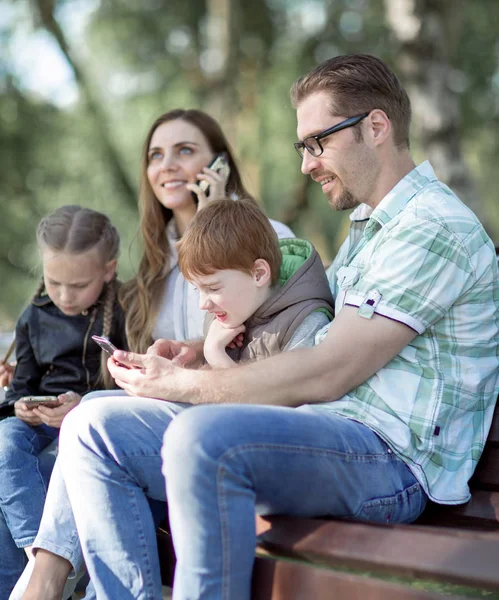 Родители и дети с помощью смартфонов сидят на скамейке в парке . — стоковое фото
