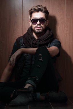 Güneş gözlüklü rahat moda adamı stüdyoda tahtaya oturur.