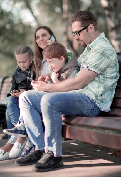 Родители и дети с помощью смартфонов сидят на скамейке в парке . — стоковое фото