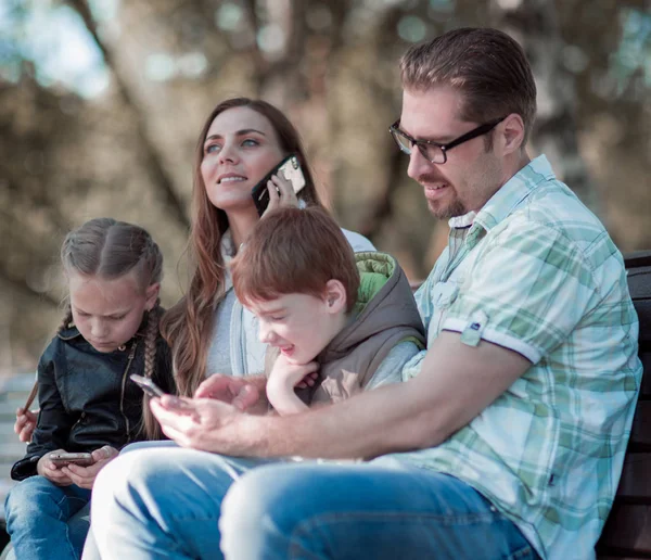 Ouders en kinderen die gebruik maken van hun smartphones zittend op een bankje. — Stockfoto