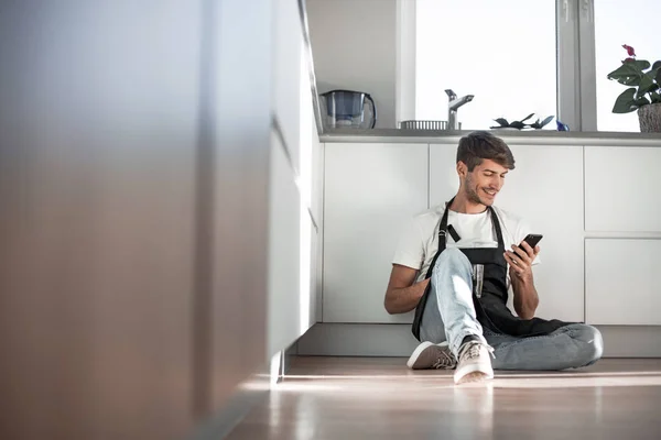 Młody człowiek ze smartfonem siedzi na podłodze w kuchni — Zdjęcie stockowe