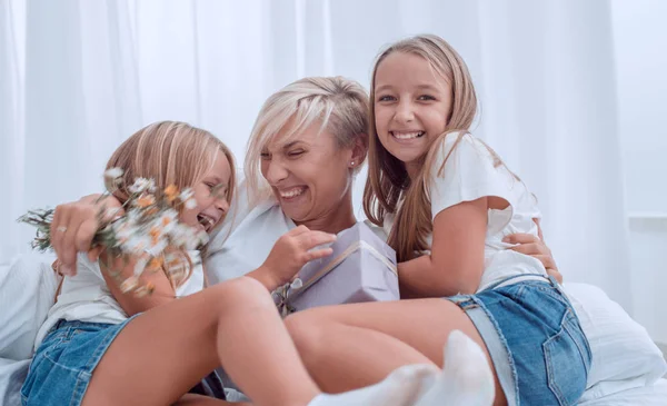 Mãe feliz com flores e presentes sentados com suas filhas na cama . — Fotografia de Stock