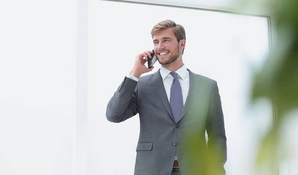 Biznesmen rozmawiający na telefon komórkowy stojący w pobliżu okna biurowego — Zdjęcie stockowe