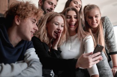 Neşeli genç adam oturmuş ve şaşırmış bir şekilde akıllı telefon kullanıyor.