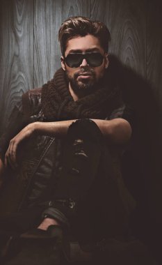 Güneş gözlüklü rahat moda adamı stüdyoda tahtaya oturur.