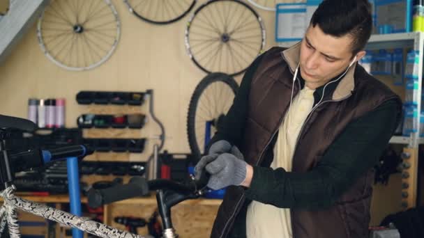 El hombre de servicio ocupado está desmontando el manillar del ciclo y limpiando piezas con tela mientras mantiene la bicicleta en el taller. Youg hombre está escuchando música con auriculares . — Vídeos de Stock