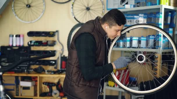 Young serviceman is cleaning bicycle wheel outside and inside with tools and piece of cloth and listening to music with earphones. Profession and people concept. — Stock Video