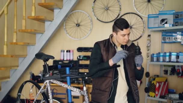 O reparador concentrado está lubrificando o mecanismo ao reparar a bicicleta na oficina agradável. Jovem de colete quente e luvas de proteção está ouvindo música  . — Vídeo de Stock