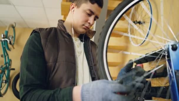 Mecánico concentrado está comprobando la rueda giratoria de la bicicleta y luego la fijación de radios con herramientas especiales durante el mantenimiento de la bicicleta y escuchar música con auriculares . — Vídeos de Stock