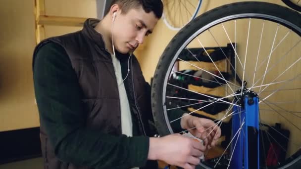 Genç adam serbest meslek makinist bisiklet tekerleği atölyede tamir ediyor. Genç adam rahat kıyafetler, sıcak yelek giyiyor ve kulaklık ile müzik dinlemek. — Stok video