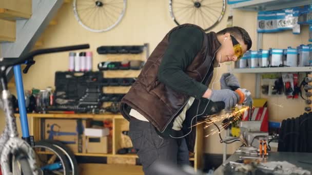 Mecánico concentrado está aserrando parte de metal con sierra circular eléctrica de pie en la mesa de trabajo en taller acogedor. Bicicletas, piezas de repuesto y equipos son visibles . — Vídeo de stock