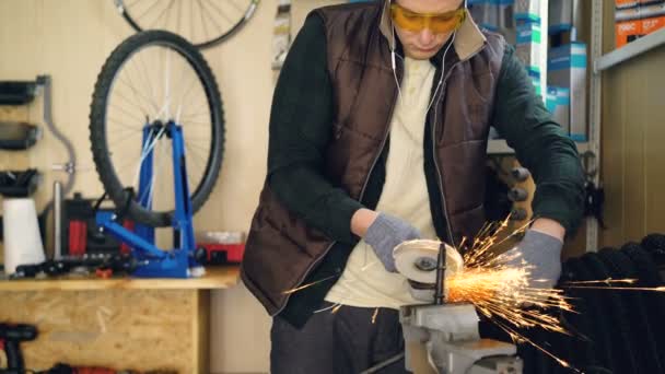 Incline-se tiro de mecânico que trabalha com serra circular elétrica que fixa a pequena parte metálica. Óculos de proteção e luvas de proteção, peças de reposição para bicicletas e ferramentas são visíveis . — Vídeo de Stock