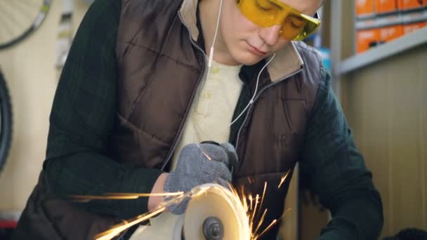 Serious repairman in safety goggles is using electric circular saw while sawing metal bicycle spare part in workplace. Bright sparks are in foreground. — Stock Video