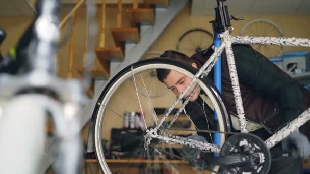 Απασχολημένος εξυπηρετητής έλεγχος πίσω τροχού περιστρεφόμενη πετάλια και κοιτάζοντας ακτίνες κατά την επισκευή των ποδηλάτων στο studio. Ωραίο εργαστήριο εσωτερικό είναι στο παρασκήνιο. — Αρχείο Βίντεο