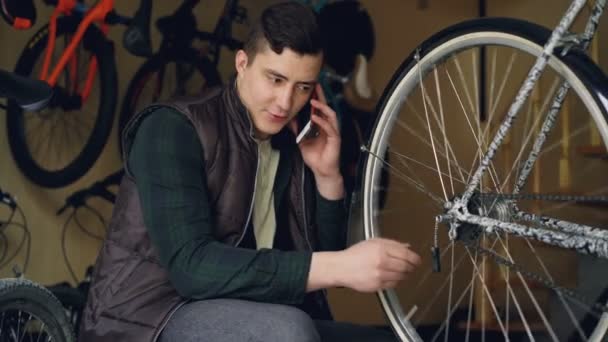 L'homme d'entretien joyeux parle sur le téléphone portable tout en travaillant dans l'atelier de rapair vélo vérifier rayons de roue et roue tournante. Concept de réparation et de communication . — Video