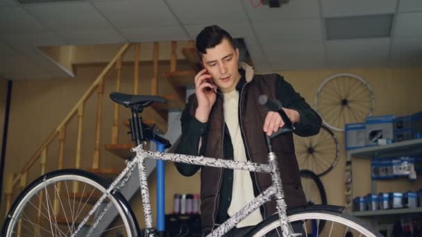 Atrakcyjny mężczyzna młody wesoły mechanik jest rozmowy na telefon komórkowy i montaż handle bar rowerów. Ludzie, konserwacji i komunikacja koncepcja. — Wideo stockowe